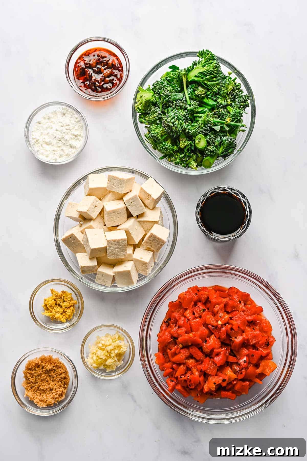 Crispy Tofu Stir-Fry 3 A vibrant display of fresh ingredients for spicy chili tofu stir fry, including pressed tofu, broccolini, roasted red peppers, garlic, ginger, soy sauce, brown sugar, and chili crunch sauce, all neatly arranged on a rustic wooden board.