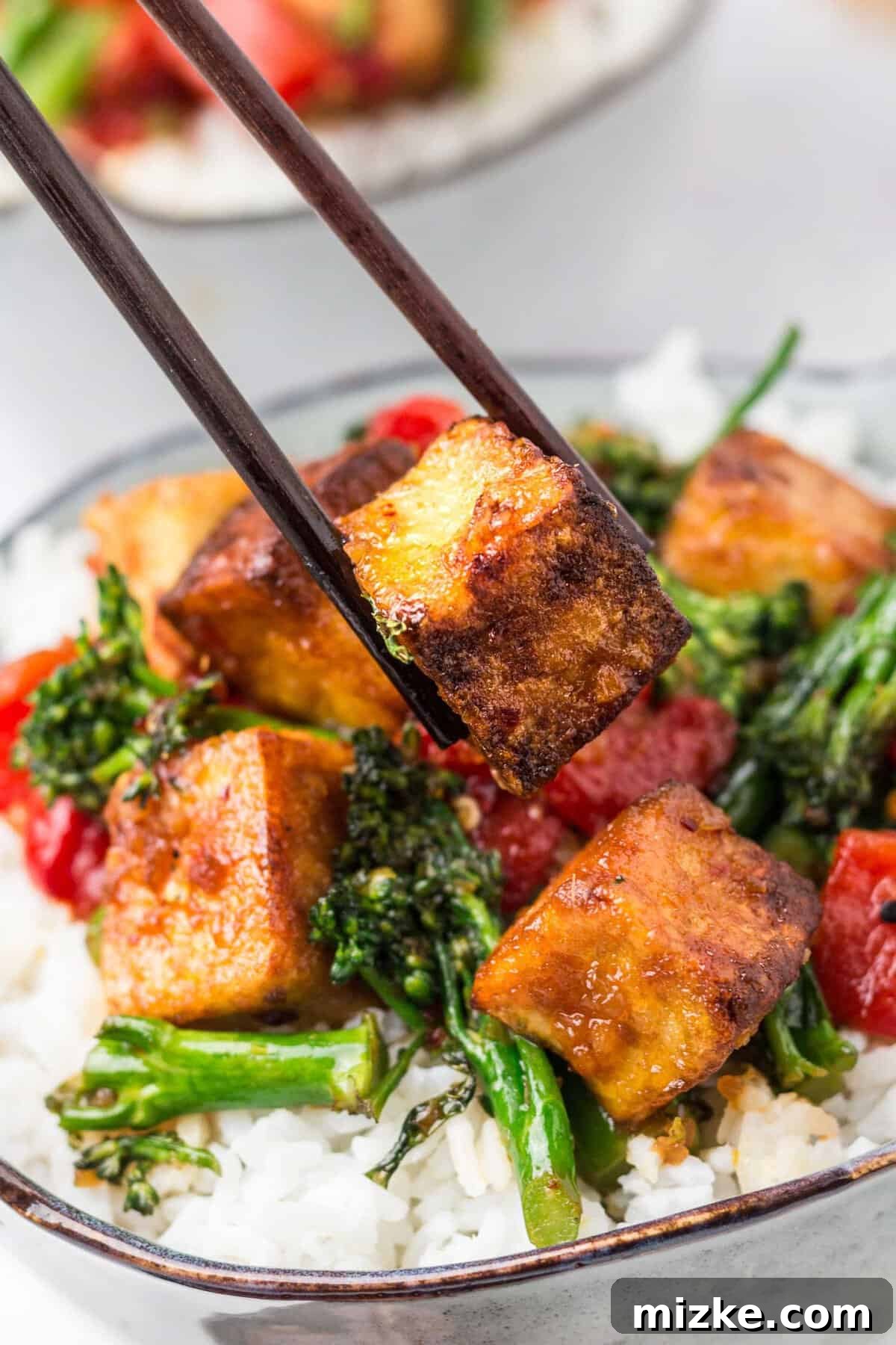 Crispy Tofu Stir-Fry 4 A glistening piece of perfectly crispy tofu is being expertly lifted from a steaming bowl of stir fry by a pair of elegant dark brown chopsticks, highlighting its golden texture and inviting appeal.