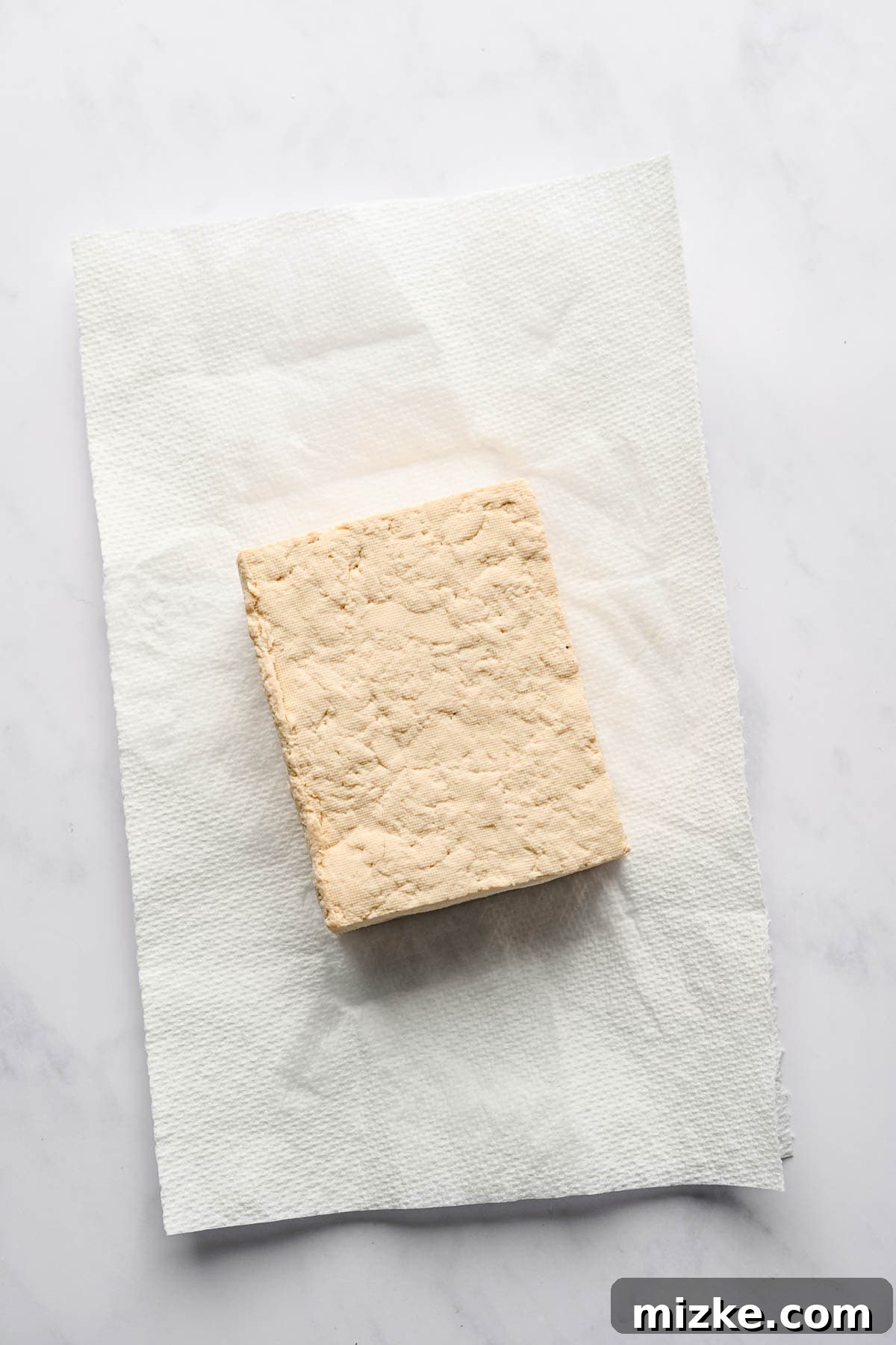 Crispy Tofu Stir-Fry 6 A dense block of extra-firm tofu rests on a stack of paper towels on a white plate, prepared for pressing to remove excess moisture.