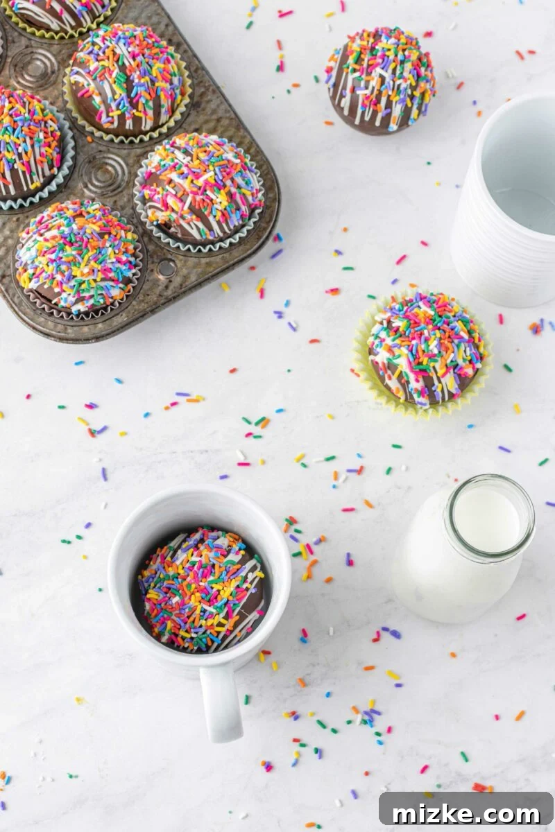 Vibrant Chocolate Dissolves 5 Sprinkles are spread out on a white countertop. A white mug contains a cocoa bomb next to a small jar of milk, across from a tray of more cocoa bombs, ready to be enjoyed.