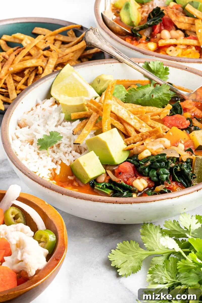 Zesty Mexican Veggie Bean Tortilla Soup 3 Cilantro and tortilla strips have been placed next to a full bowl of Mexican soup on a white surface.