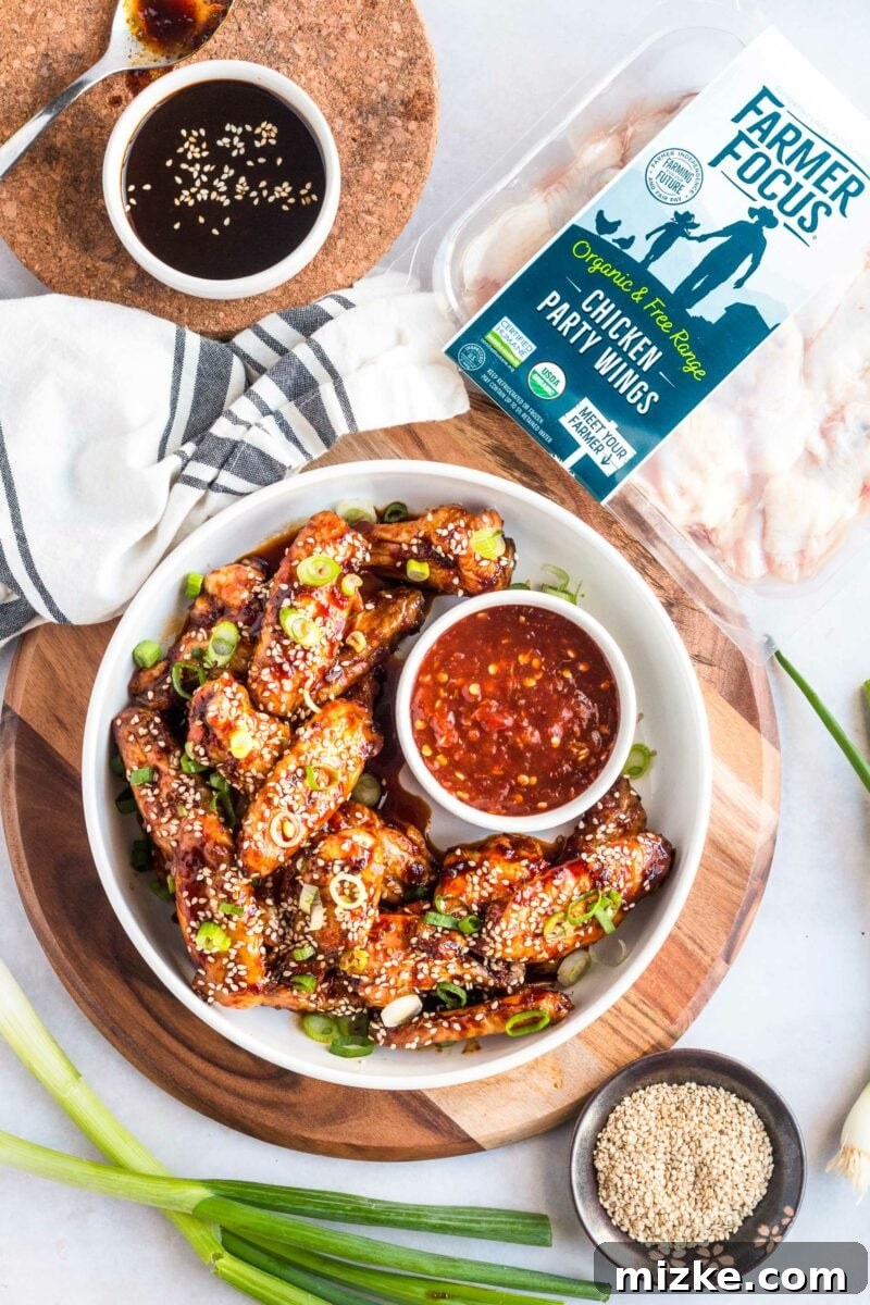 Gochujang Glazed Wings 2 A bowl of ready-to-eat sticky Korean chicken wings garnished with sesame seeds and green onions, positioned next to an unopened package of Farmer Focus chicken party wings.