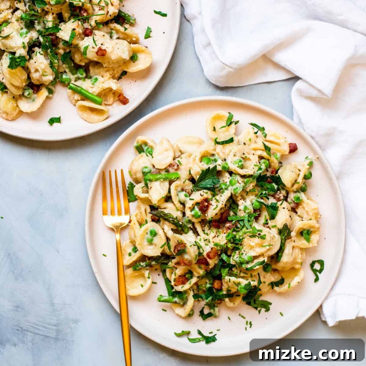 Two plates full of creamy pasta with asparagus, peas, and pancetta have been placed next to one another on a white surface.