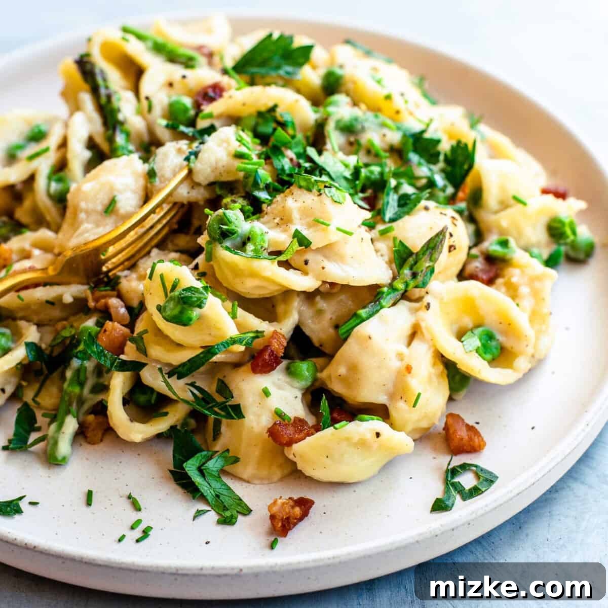 Fresh herbs garnish a serving of creamy noodles, asparagus, peas and crispy pancetta on a white plate.