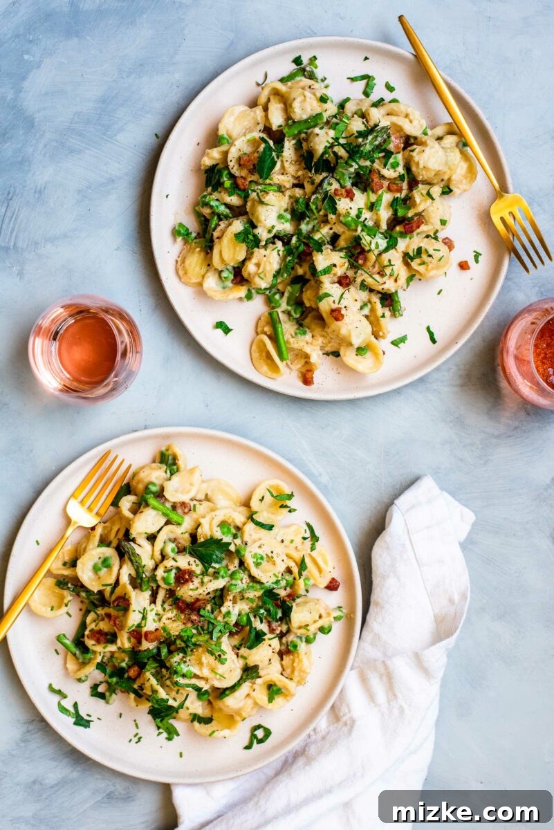 Two plates of pasta with asparagus, peas, and pancetta are placed next to two glasses of rosé.