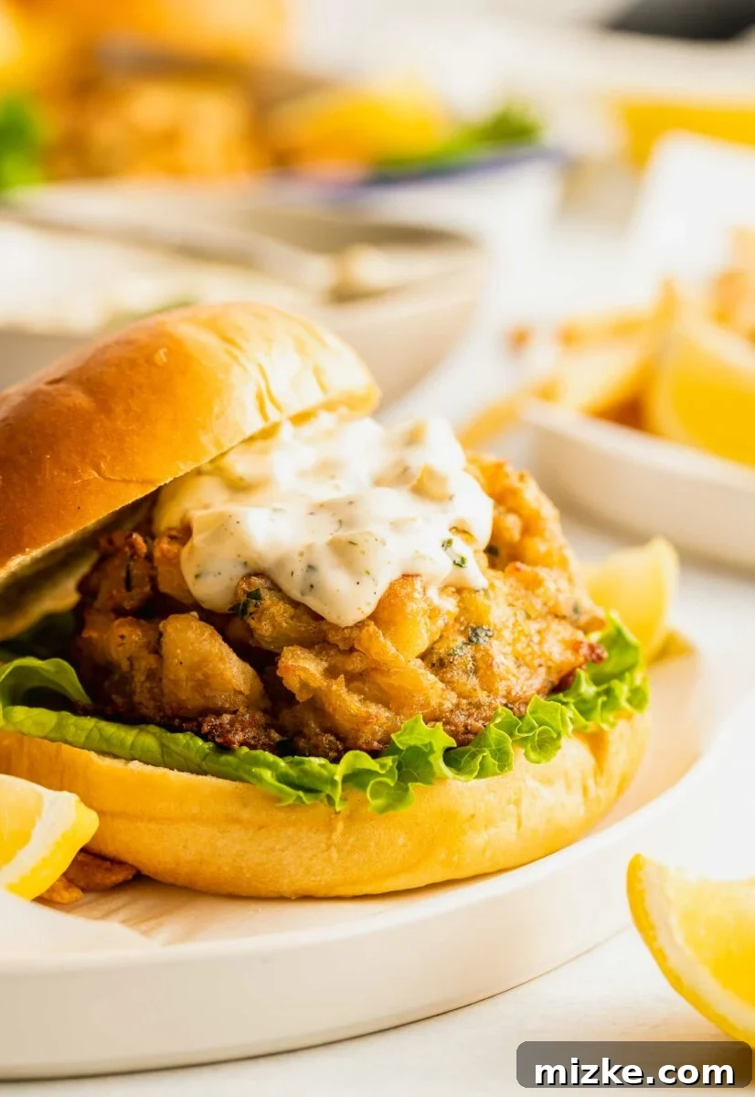Ultimate Crab Cake Sandwiches 6 A perfectly constructed crab cake sandwich reveals its delicious layers: a generous dollop of creamy tartar sauce, crisp green lettuce, and a succulent crab cake, all nestled within a soft bun.