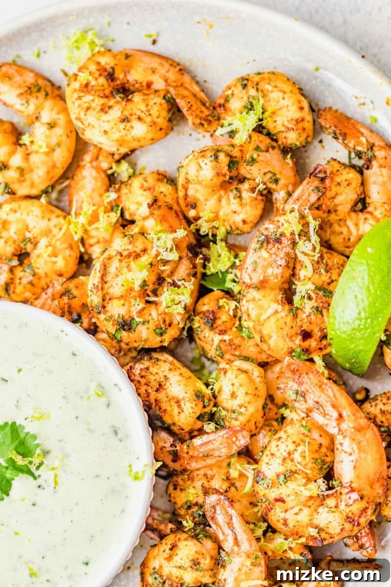 A large plate of vibrant, air-fried spicy cilantro lime shrimp is presented next to a small bowl of creamy green cilantro lime dressing, garnished with fresh herbs and lime wedges, ready to be served as an appetizer.