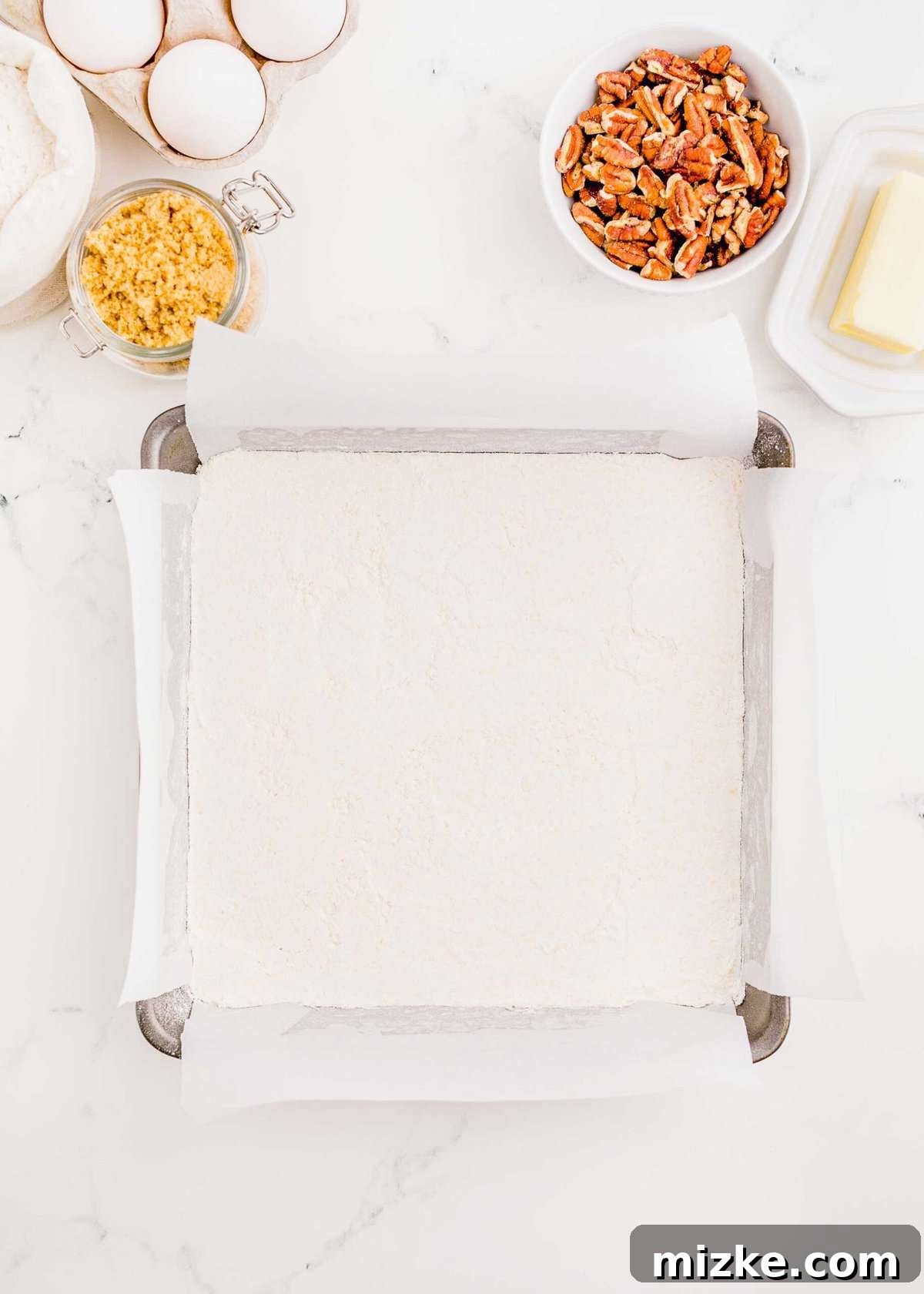 Golden Pecan Pie Squares 7 The crumbly shortbread crust mixture has been evenly pressed into the bottom of a 9-inch square baking sheet lined with parchment paper, forming a solid base for the pecan filling.