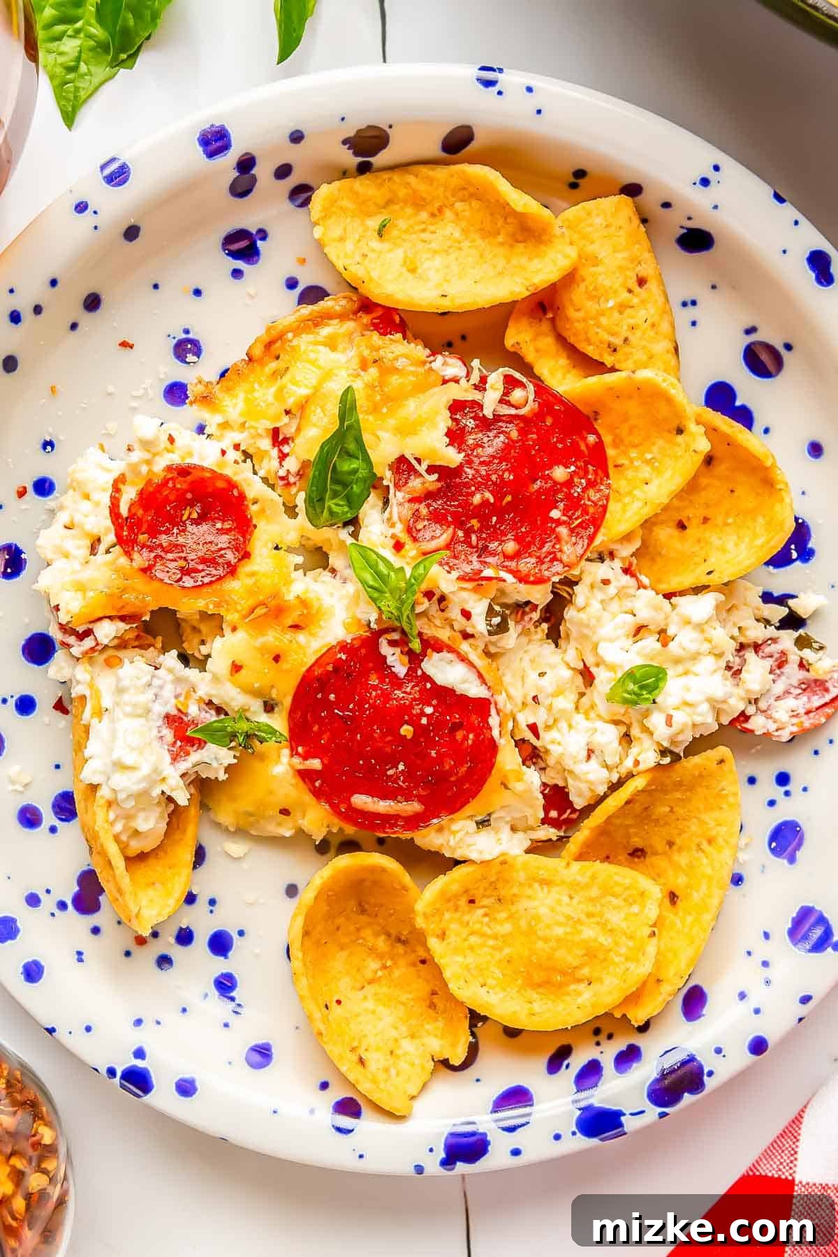 Cheesy Pepperoni Pizza Bake 4 A serving of pepperoni pizza dip on a speckled white and blue plate, garnished with fresh basil, next to a small bowl of corn tortilla scoops