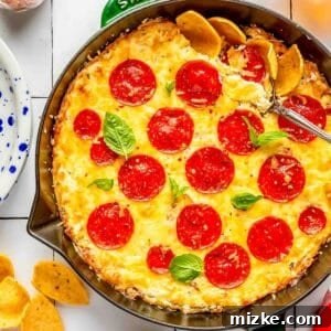 Pepperoni pizza dip in a green cast iron skillet with a metal spoon in the corner, showing a close-up of the bubbly cheese