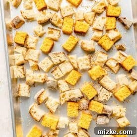 Cubed cornbread and sourdough bread spread on a metal baking sheet, ready for toasting in the oven.