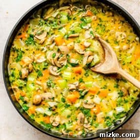 Sautéed vegetables and chicken broth simmering in a large skillet with a wooden spoon.