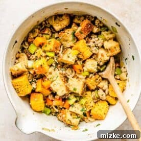 Toasted cornbread and sourdough cubes mixed with the savory vegetable and sausage mixture in a large heat-proof mixing bowl, being gently tossed with a wooden spoon.