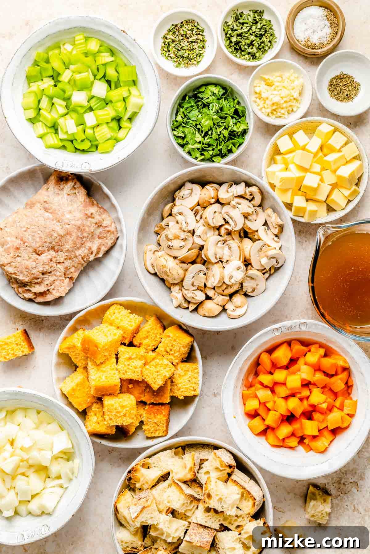 Various fresh ingredients for cornbread stuffing laid out on a white surface, including sourdough bread, cornbread, fresh herbs, vegetables, and sausage.