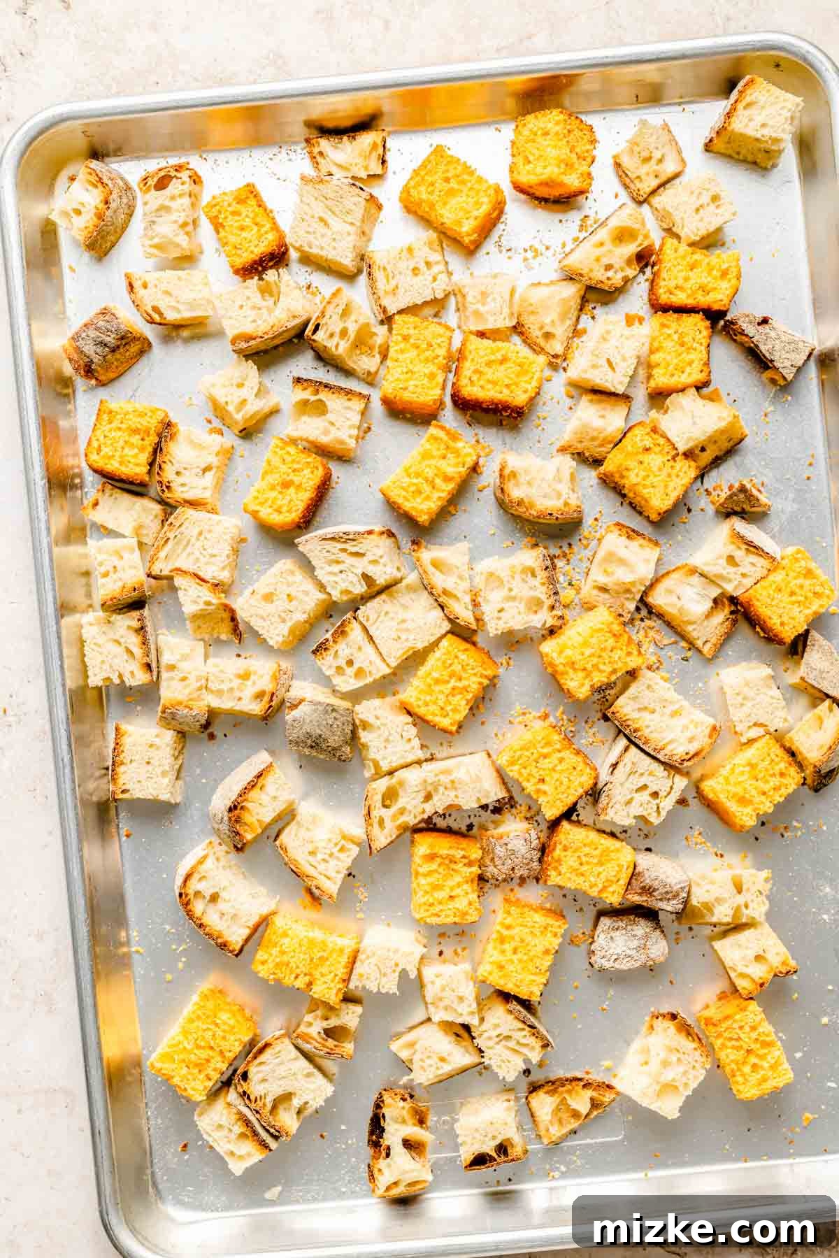 Cubed cornbread and sourdough bread spread on a metal baking sheet, ready for toasting in the oven.
