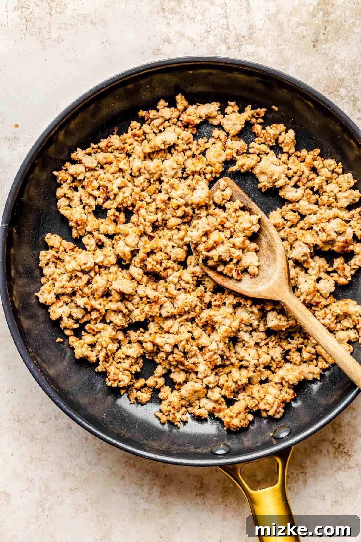 Crumbled breakfast sausage browning in a skillet with a wooden spoon, releasing savory aromas.
