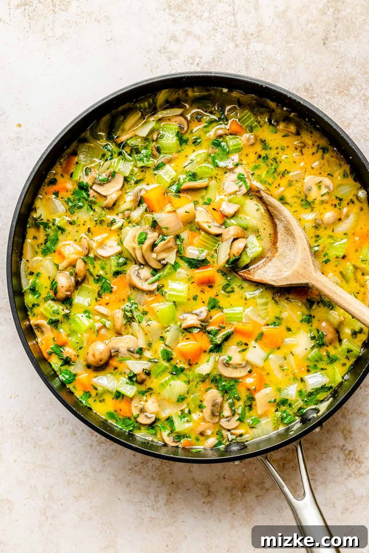 Sautéed vegetables and chicken broth simmering in a large skillet with a wooden spoon.