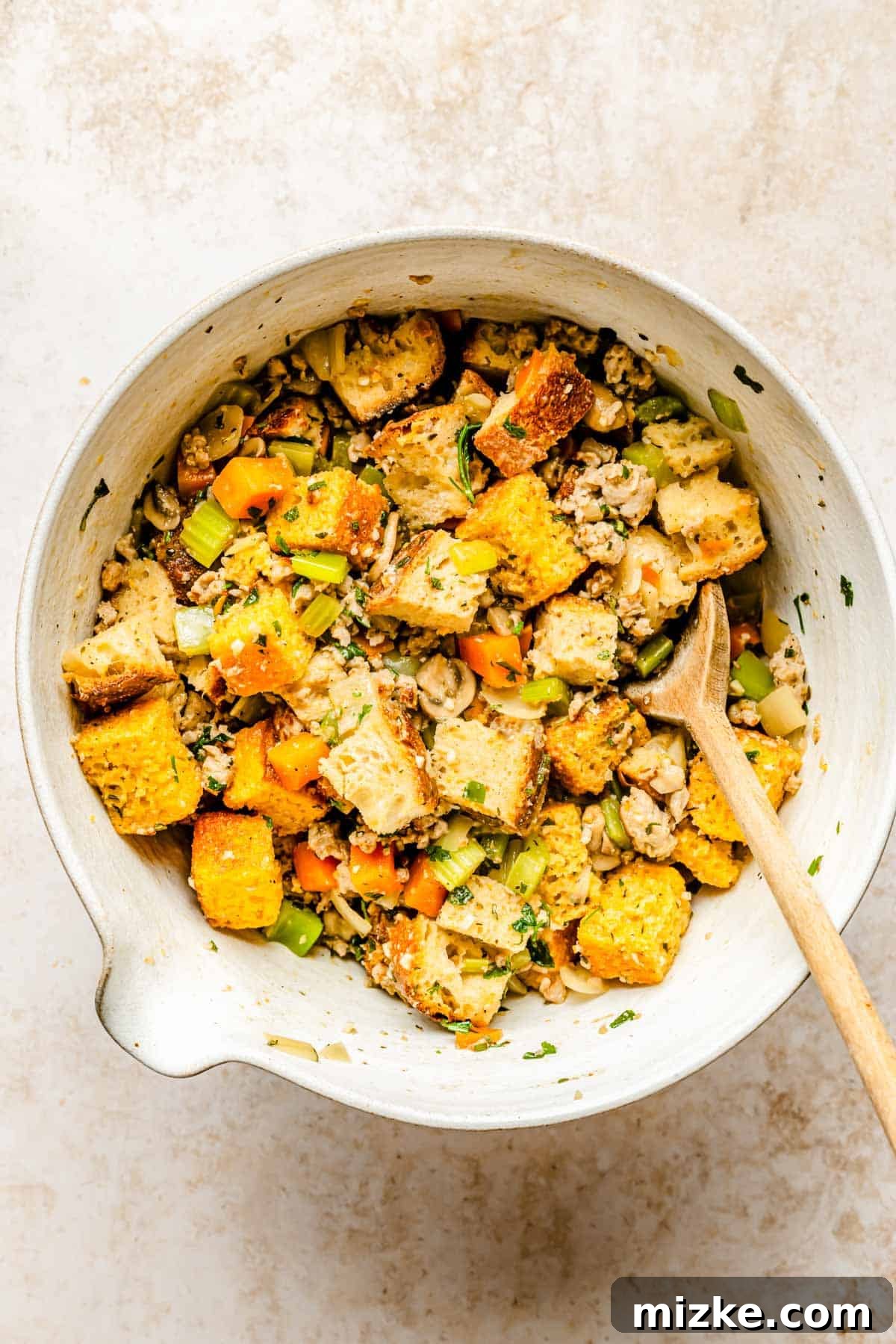 Toasted cornbread and sourdough cubes mixed with the savory vegetable and sausage mixture in a large heat-proof mixing bowl, being gently tossed with a wooden spoon.