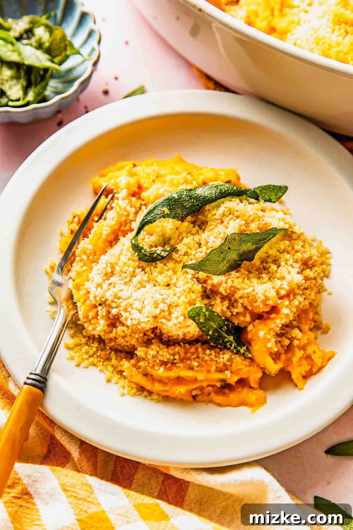 Rustic Savory Sweet Potato Bake 3 A plate of savory sweet potato casserole filling topped with breadcrumbs and crispy sage leaves, served with a fork on a white plate.