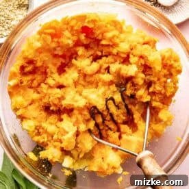 Rustic Savory Sweet Potato Bake 6 A glass bowl of mashed potatoes with a potato masher, surrounded by spices, salt, pepper, and fresh sage leaves on a countertop.