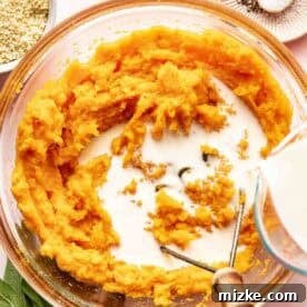 Rustic Savory Sweet Potato Bake 8 A bowl of mashed sweet potatoes with milk being poured in, surrounded by a bowl of breadcrumbs, fresh sage, and a plate of salt and pepper.