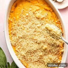 Rustic Savory Sweet Potato Bake 9 An oval white baking dish filled with sweet potato casserole topped with breadcrumb mixture; spoon adding more crumbs. A plate with salt and pepper and fresh sage leaves nearby.