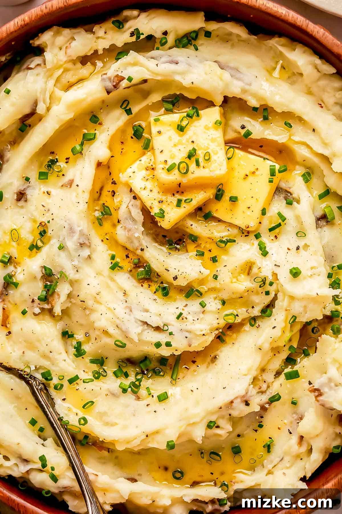 Heavenly Garlic Mashed Potatoes 3 An appetizing close-up of creamy garlic mashed potatoes, showcasing their smooth texture with three generous pats of butter gently melting into the surface, garnished with fresh green chives. A metal spoon rests nearby, inviting a taste.
