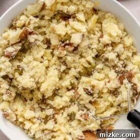 Heavenly Garlic Mashed Potatoes 7 Boiled potatoes being mashed in a white mixing bowl using a potato masher, aiming for a smooth consistency.