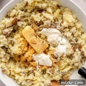 Heavenly Garlic Mashed Potatoes 8 Butter, sour cream, garlic powder, salt, and pepper being added to the mashed potatoes in a white bowl, ready for mixing.