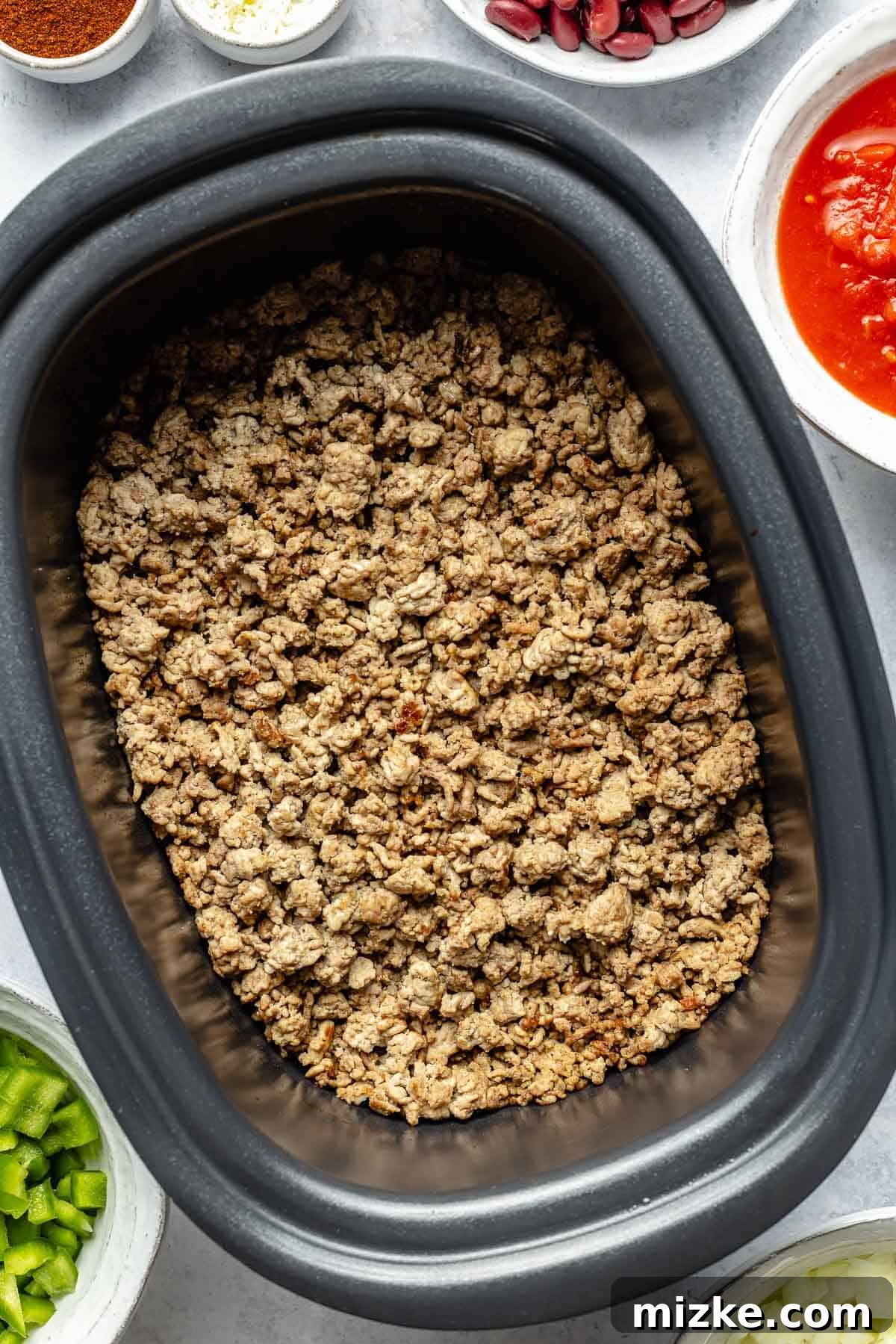 The first step: browning the meat before adding it to the slow cooker for a rich chili. Close-up shot of browned ground meat being added to the slow cooker insert, ready for the next steps of making chili.