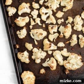 Roasted cauliflower florets spread on a metal baking pan, ready to be tossed in buffalo sauce.