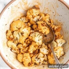 Roasted cauliflower being tossed with buffalo sauce in a white ceramic bowl using a metal spoon.