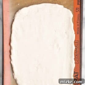 Pizza dough pressed into a rectangular shape on a silicone baking mat on a baking sheet.