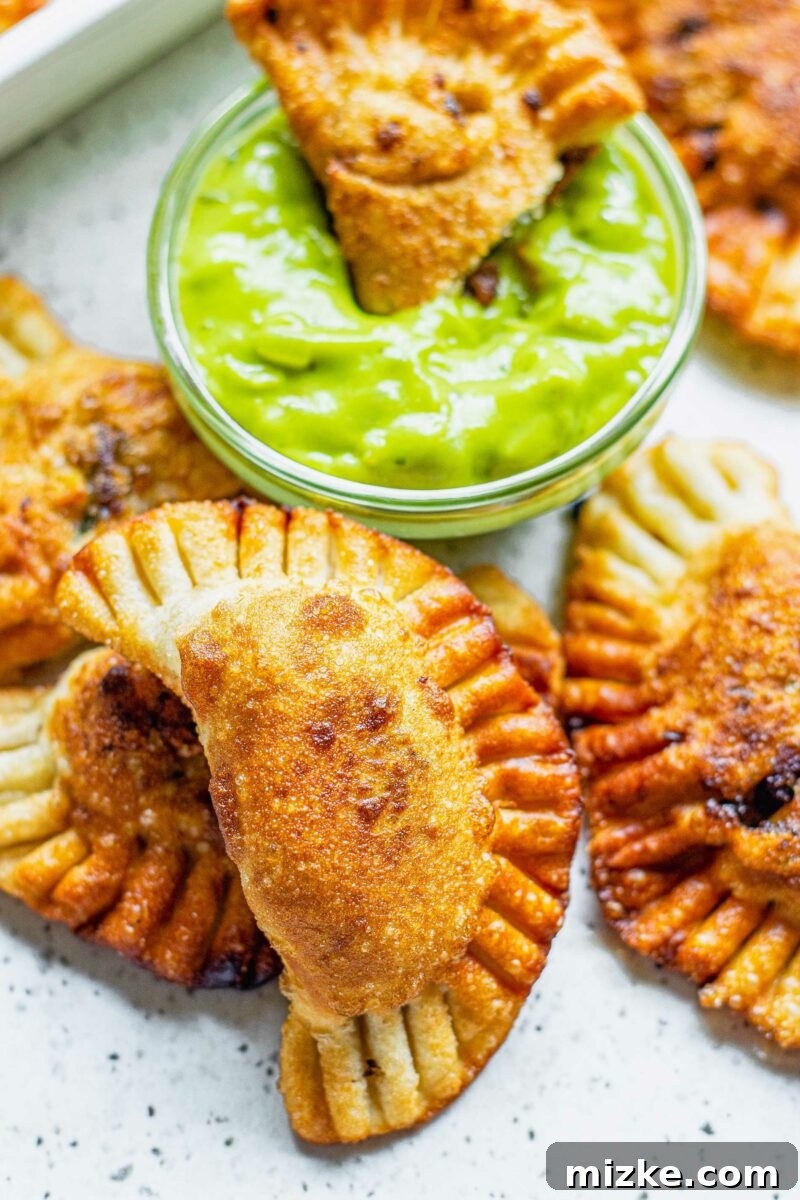 A freshly fried beef empanada is being carefully dipped into a small bowl of vibrant green sauce, highlighting its golden crust and delicious filling.