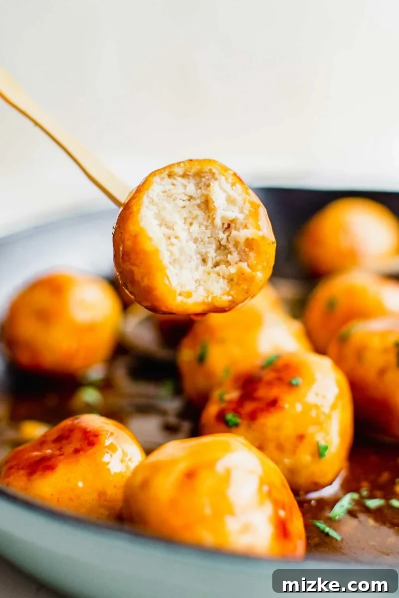 Fiery Honey Chicken Meatballs 3 A skewer is lifting a perfectly cooked, bitten-into chicken meatball from a dark pan, showcasing its juicy interior and sticky hot honey coating.
