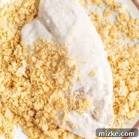 Tilapia coated in batter being dipped into a plate of crushed tortilla chips.