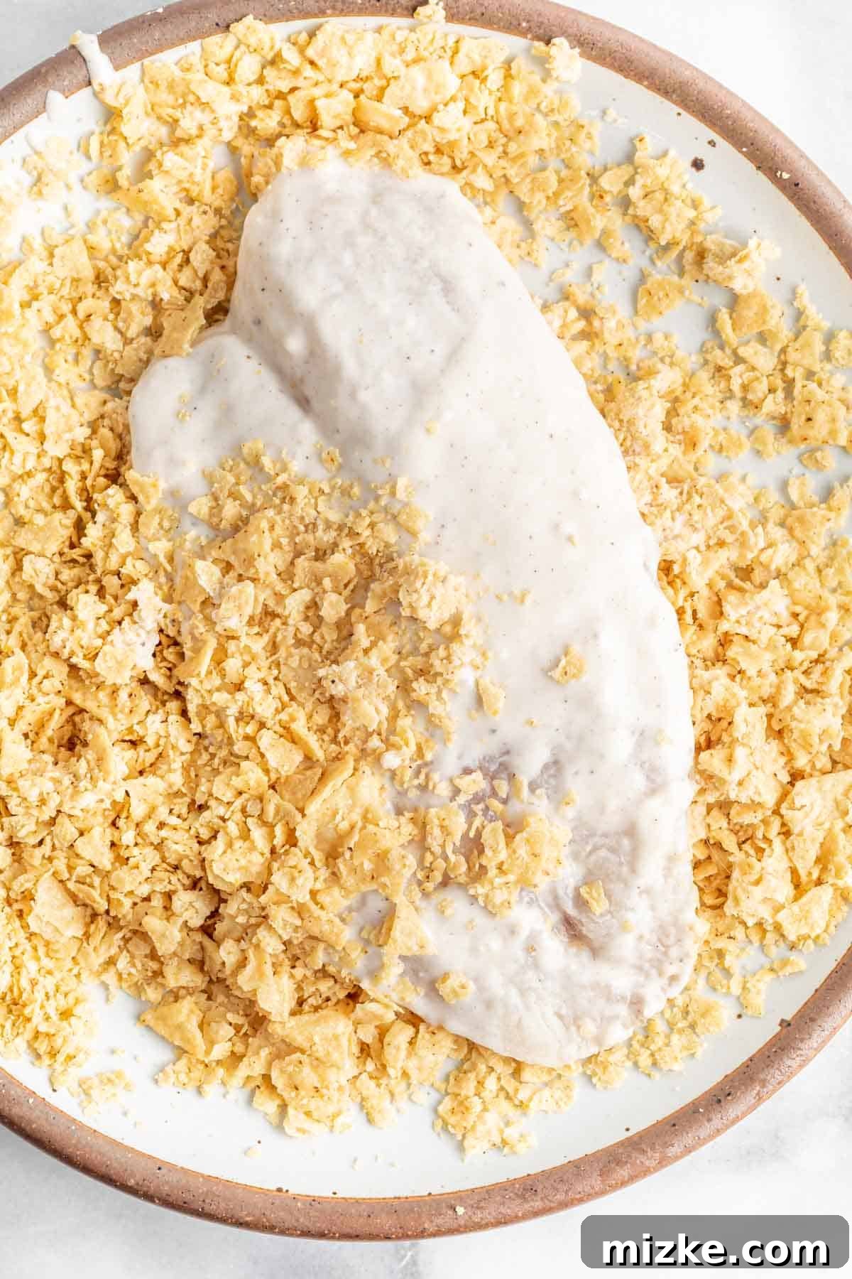 Southwestern Tilapia Crunch 6 A batter-coated tilapia fillet is being gently pressed into a plate of crushed tortilla chips to create a crisp crust.