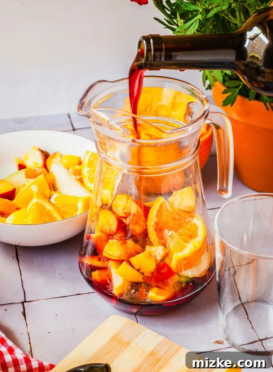 Red wine is being poured into a pitcher with many sliced fruits. 