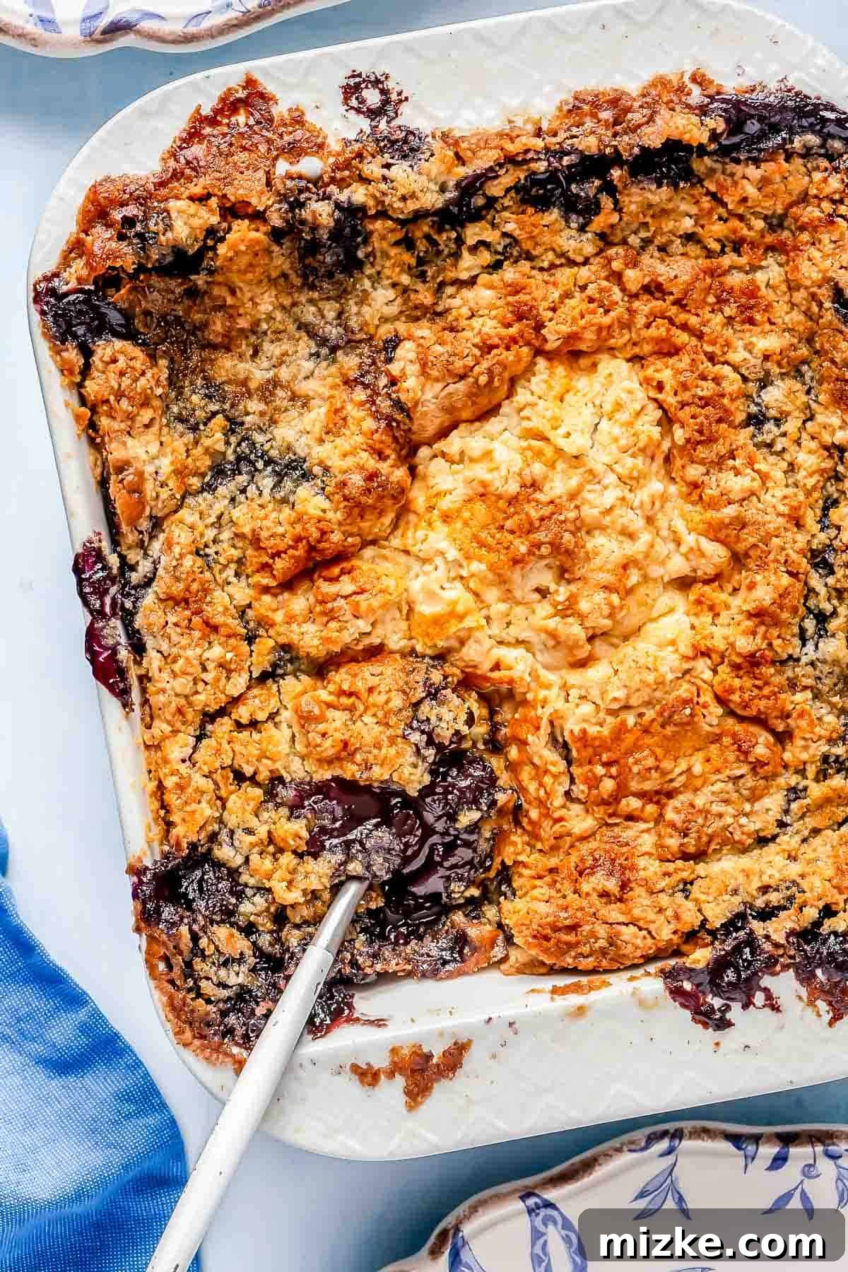 Baked Blueberry Dump Cake A beautifully baked blueberry dump cake in a square white baking pan, showcasing a golden-brown, crispy topping with a metal spoon resting in the center. The cake is warm and inviting.