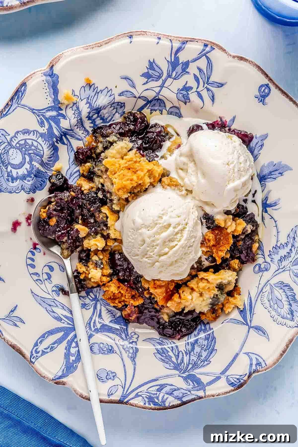 Blueberry Dump Cake with Melting Vanilla Ice Cream Two delightful scoops of creamy vanilla ice cream beginning to melt over a warm slice of blueberry dump cake, elegantly presented on a vibrant blue china plate.