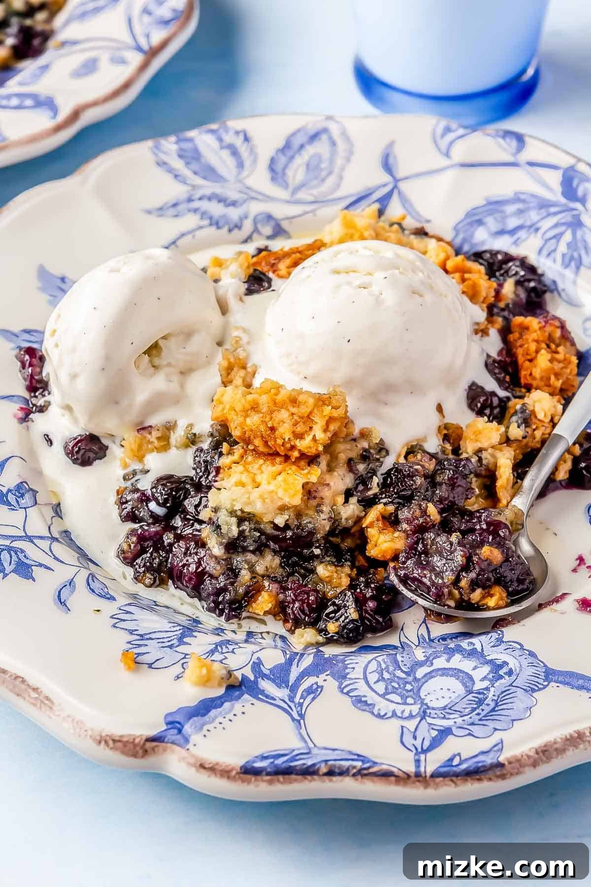 Melted Ice Cream on Blueberry Dump Cake Slice Two scoops of creamy, slightly melted vanilla ice cream beautifully topping a warm slice of blueberry dump cake. The dessert is presented on an elegant blue china plate, with a metal spoon beside it, inviting indulgence.
