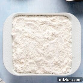 Dry cake mix is carefully poured and spread over the prepared blueberry layer in the white square baking dish, forming the next crucial layer.