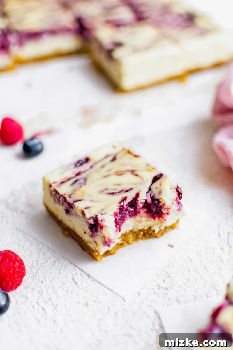 Vibrant Berry Cheesecake Squares 6 A Triple Berry Cheesecake Bar, partially eaten, rests on a small piece of parchment paper, showcasing the rich texture and berry inclusions.