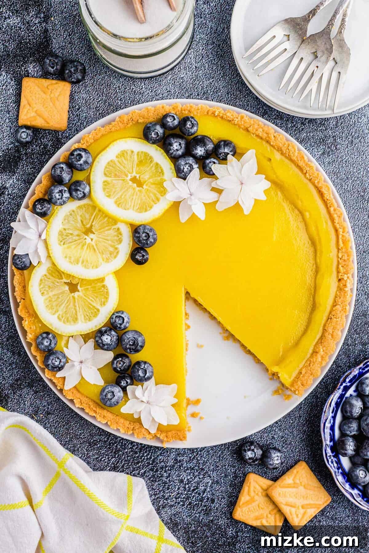 Bright Lemon Tart 2 A vibrant yellow lemon tart, elegantly garnished with fresh lemon slices, delicate white flowers, and plump blueberries, rests on a pristine white surface, ready to be served.