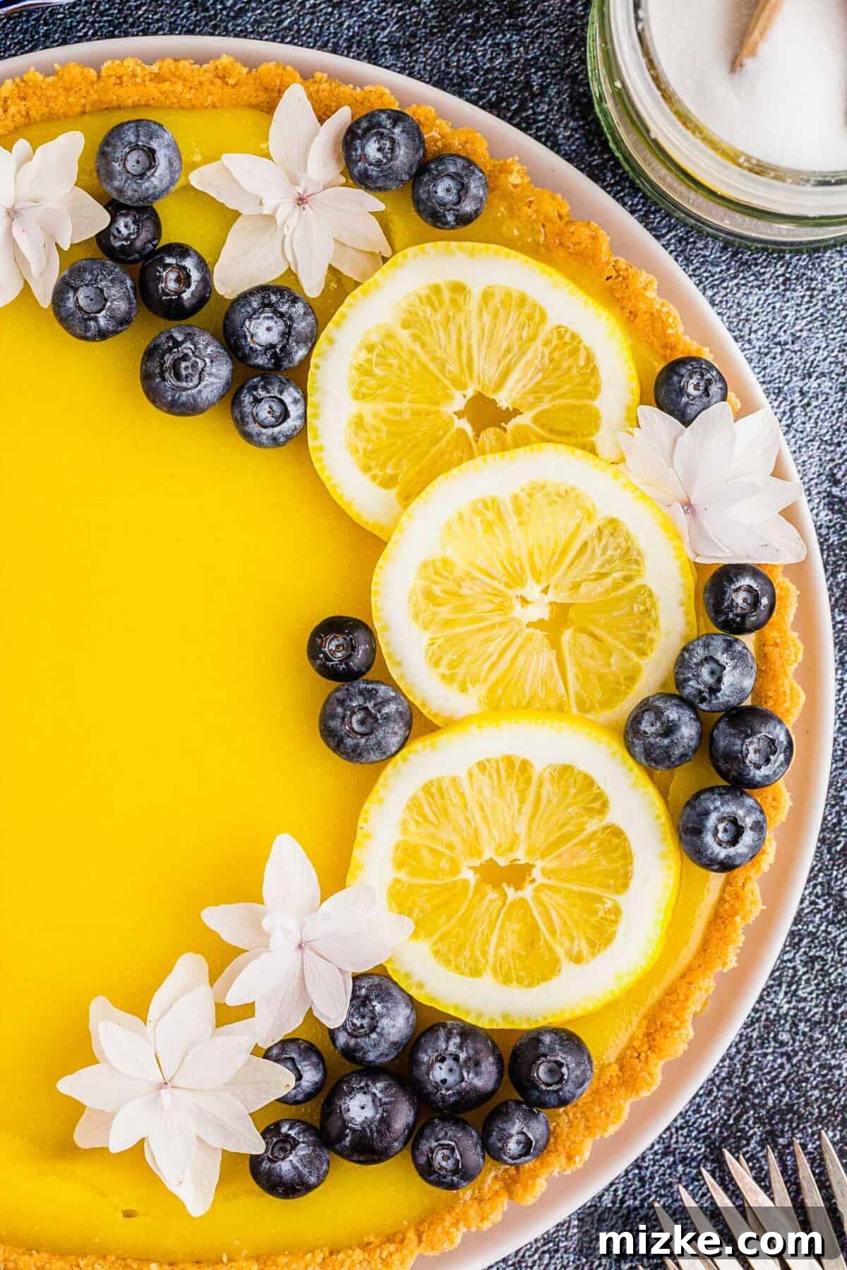Bright Lemon Tart 10 A close-up shot of a beautifully prepared lemon tart, adorned with fresh lemon slices, vibrant blueberries, and delicate white edible flowers, exquisitely arranged and ready for serving as a centerpiece dessert.