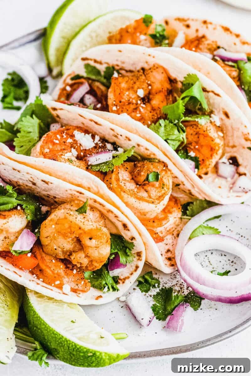 Several shrimp tacos are placed on a white surface, garnished with fresh cilantro and lime wedges.