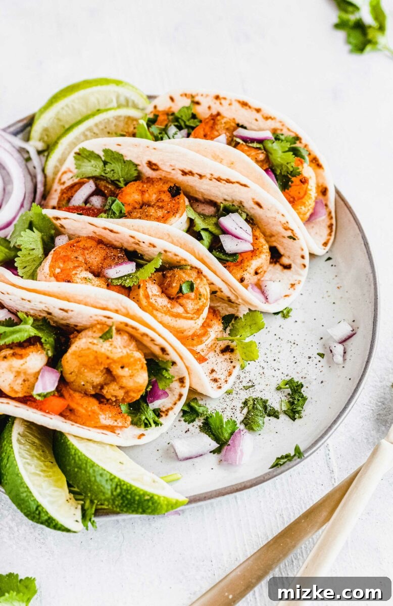 Several shrimp tacos artfully arranged on a white plate, garnished with fresh cilantro and lime wedges.