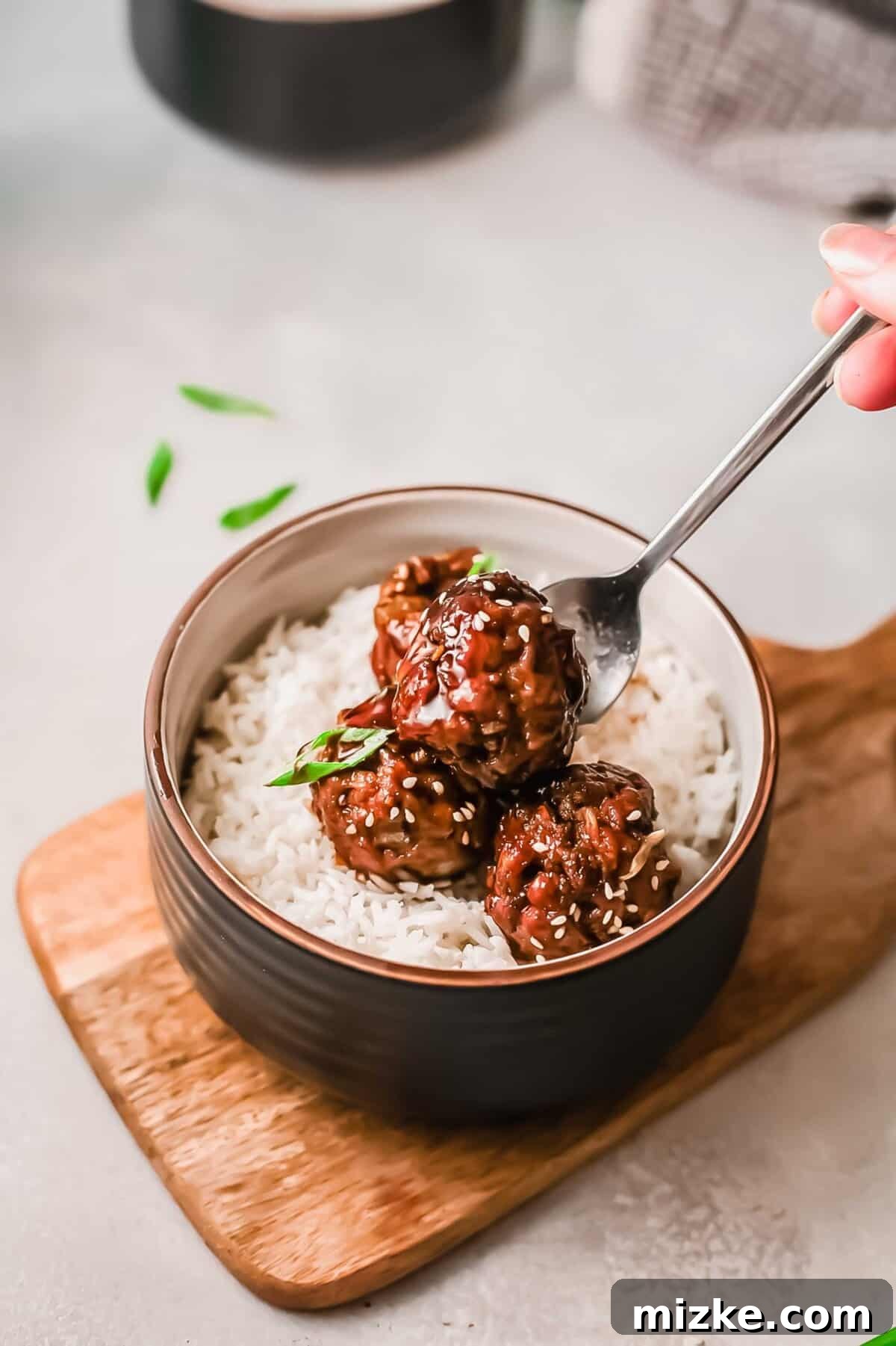 Steppe-Style Meatballs 3 A single Mongolian meatball held by a fork, hovering above a bowl of white rice on a wooden cutting board, showcasing its sticky glaze.
