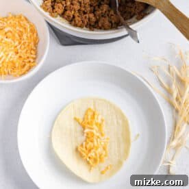 Crispy Air Fryer Beef Taquitos 15 shredded cheese on a corn tortilla on top of a corn husk string on a white plate next to a bowl of ground beef