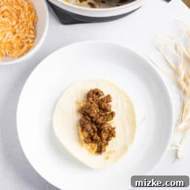 Crispy Air Fryer Beef Taquitos 16 ground beef on top of shredded cheese on a corn tortilla on top of a corn husk string on a white plate next to a bowl of ground beef